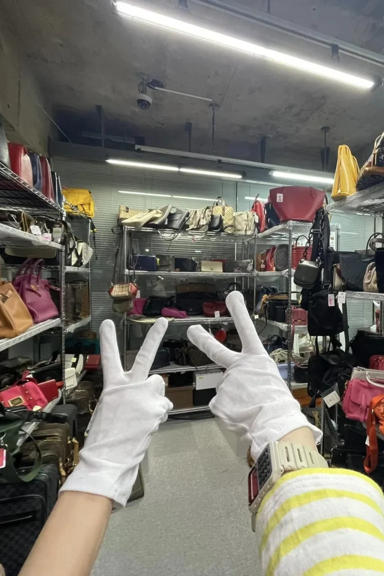 Rayonnages remplis de sacs à main de seconde main dans l'entrepôt de Tokyo, avec deux mains gantées de blanc qui font le signe V au premier plan.
