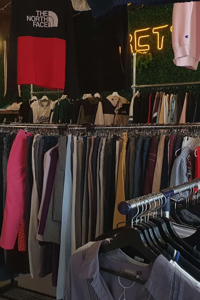 Portants de vêtements de seconde main dans la boutique EGRETTA, sous un néon jaune et un t-shirt The North Face suspendu.