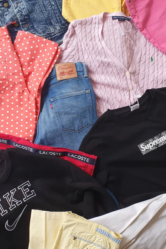 Vêtements de seconde main posés à plat : veste en jean, pull rose torsadé, chemise à pois, jean, sweats noirs et pantalon jaune pâle.