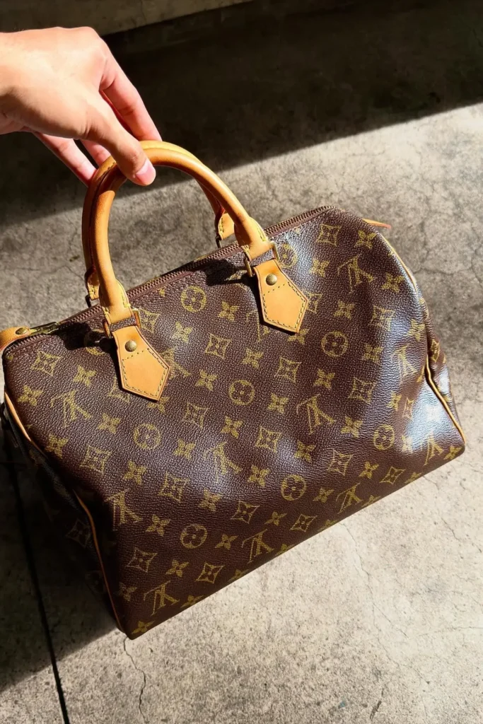 Main tenant par ses anses un sac à main Louis Vuitton en toile Monogram de seconde main, au soleil sur un sol en béton.