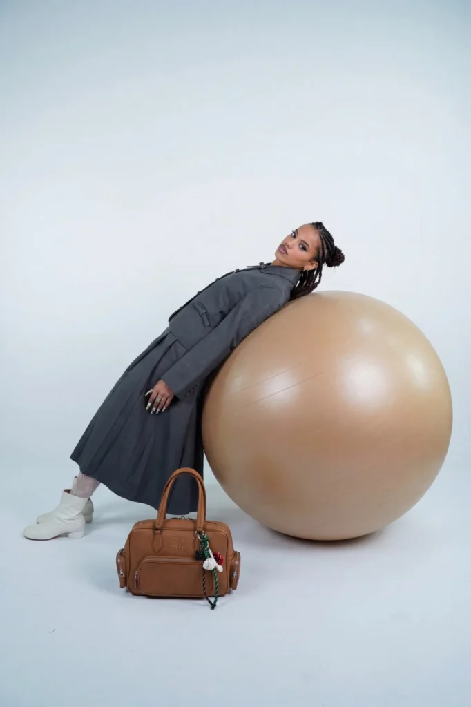 Modèle adossée à un grand ballon beige, en veste et jupe plissée grises, bottines blanches, avec un sac en cuir camel Miu Miu posé au premier plan.