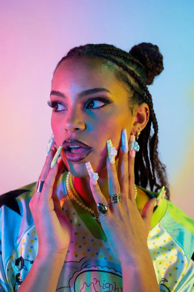 Portrait d'une modèle aux tresses relevées en chignon, maillot satiné multicolore, collier de perles et longs ongles pastel décorés, sur fond rose et bleu.