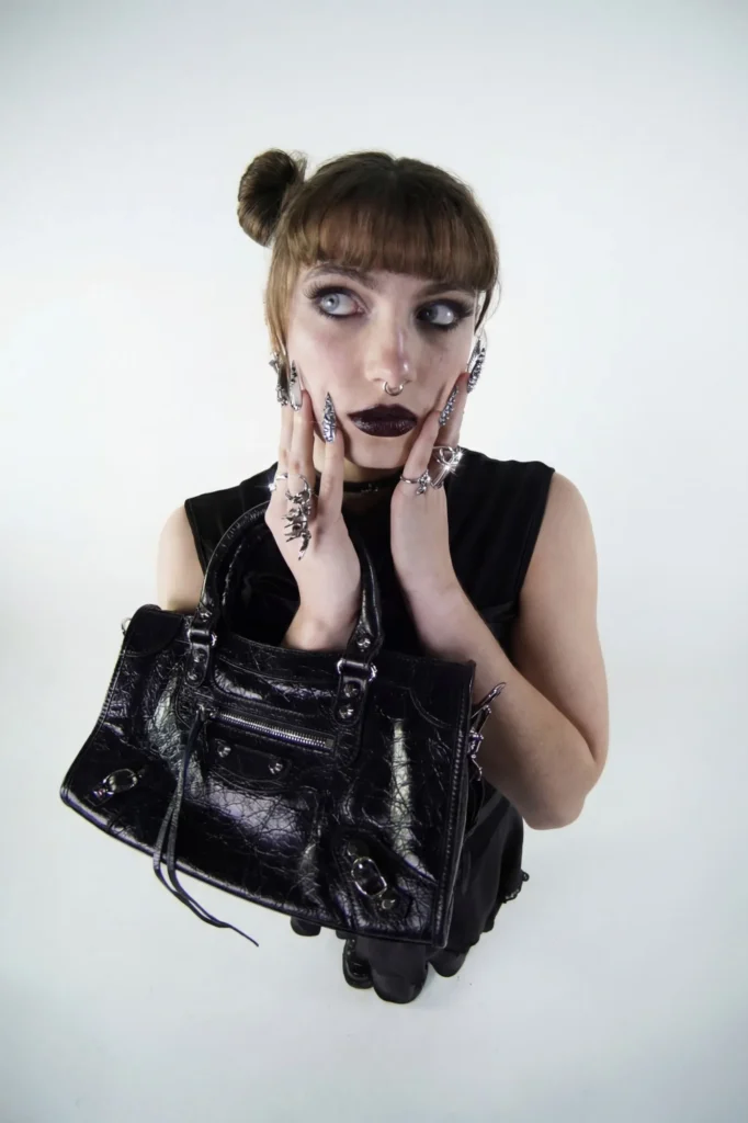 Modèle vue en plongée, tenue noire sans manches, rouge à lèvres foncé et bagues argentées, tenant un sac à main en cuir noir clouté.