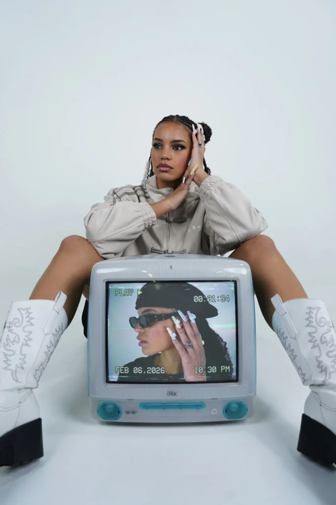 Modèle assise derrière un ancien iMac bleu, en veste adidas beige à boutons brandebourgs et bottes blanches à plateforme.
