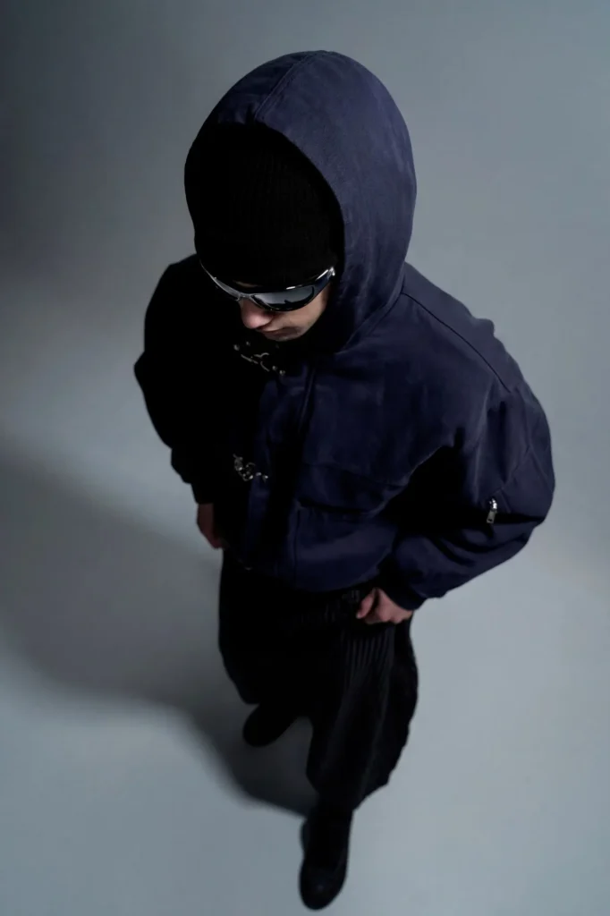 Silhouette vue de dessus en veste à capuche bleu nuit, bonnet noir, lunettes miroir et pantalon noir plissé.
