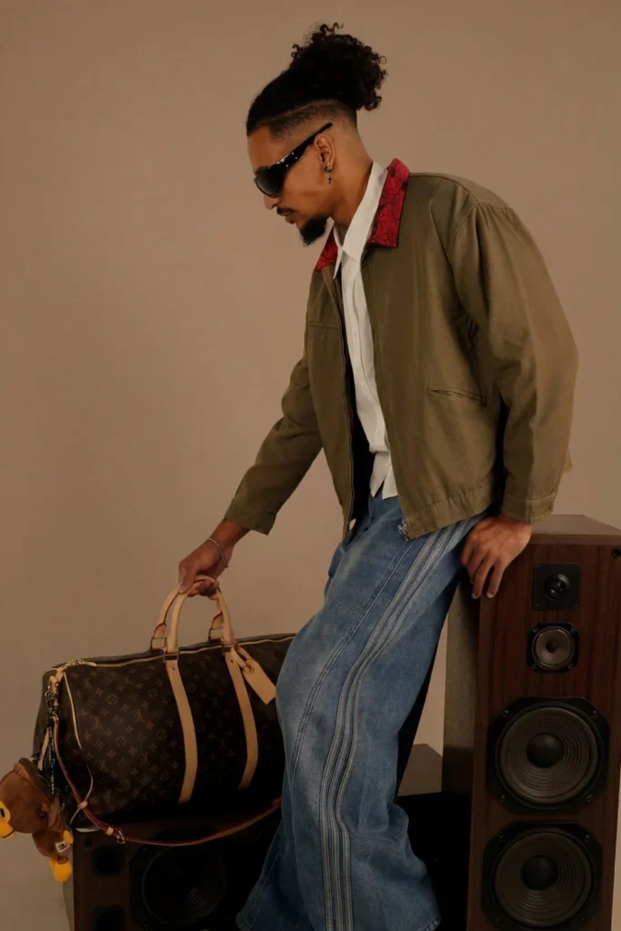 Modèle de profil en veste kaki à col rouge et jean large, tenant un sac de voyage Louis Vuitton en toile Monogram posé sur des enceintes en bois.