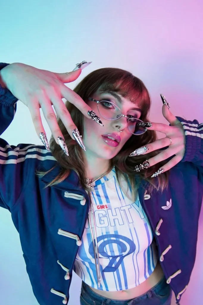 Modèle en veste de survêtement adidas bleu marine à boutons brandebourgs, haut rayé bleu et blanc, lunettes fines et longs ongles argentés, sur fond rose et bleu.